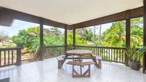 Une terrasse couverte, dotée d’une table en bois et de bancs, entourée d’une balustrade et offrant une vue sur un jardin luxuriant.
