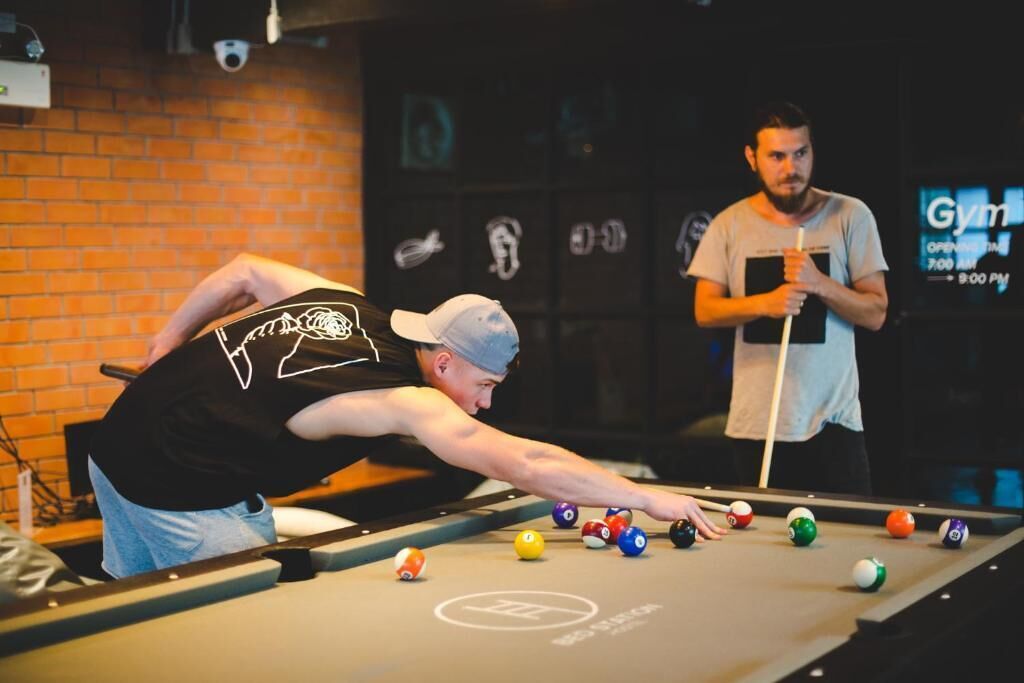 A pool table with a person playing, another person observing, and a sign indicating the gym's operating hours.