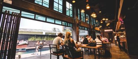 An indoor dining area with high ceilings, large windows, and hanging pendant lights. Tables are arranged for groups of people.