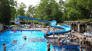 A swimming pool with a water slide, surrounded by trees and shaded areas with lounge chairs.