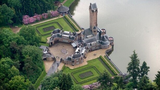 An aerial view of a historic castle with a large tower, surrounded by formal gardens and a lake.