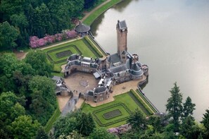 Vista aérea de un castillo histórico con una torre alta, rodeado de jardines formales y un lago.