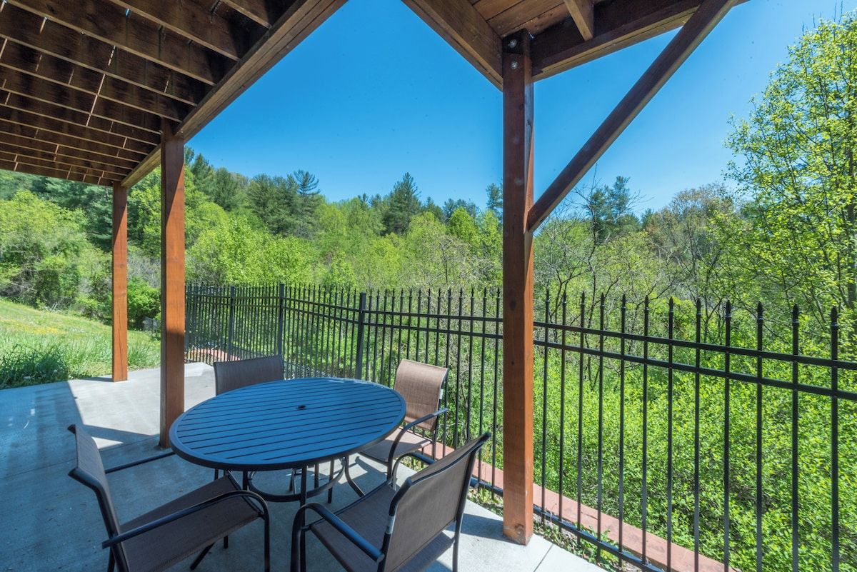 A covered outdoor seating area with a table and chairs, surrounded by a metal fence, overlooking a lush green landscape.