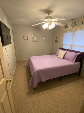 Un dormitorio con cama, ventilador de techo, ventana con persianas y puerta con picaporte.