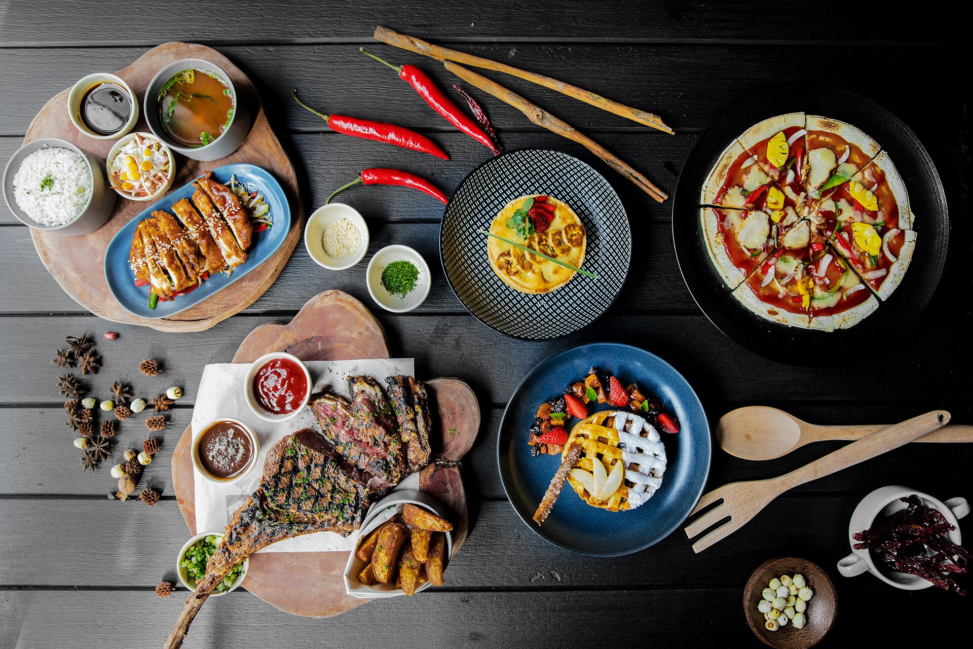 Variedad de platos, incluyendo carne a la parrilla, arroz y una pizza, dispuestos sobre una mesa de madera.