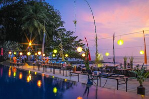 An outdoor pool area with tables and chairs, palm trees, and a view of the ocean at sunset. - Mambruk Hotel & Convention (Anyer)