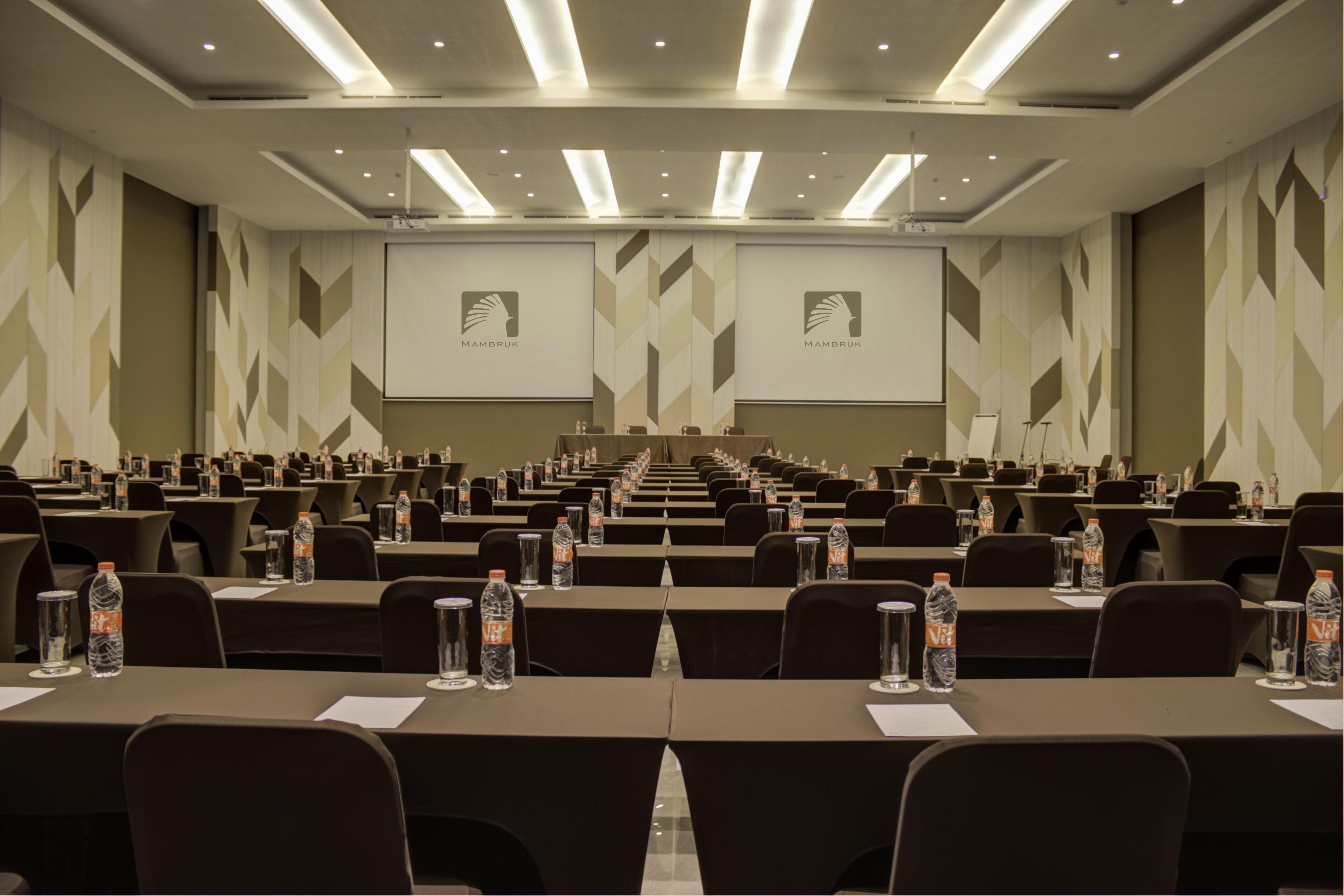 A conference room with rows of tables, water bottles, and presentation screens.
