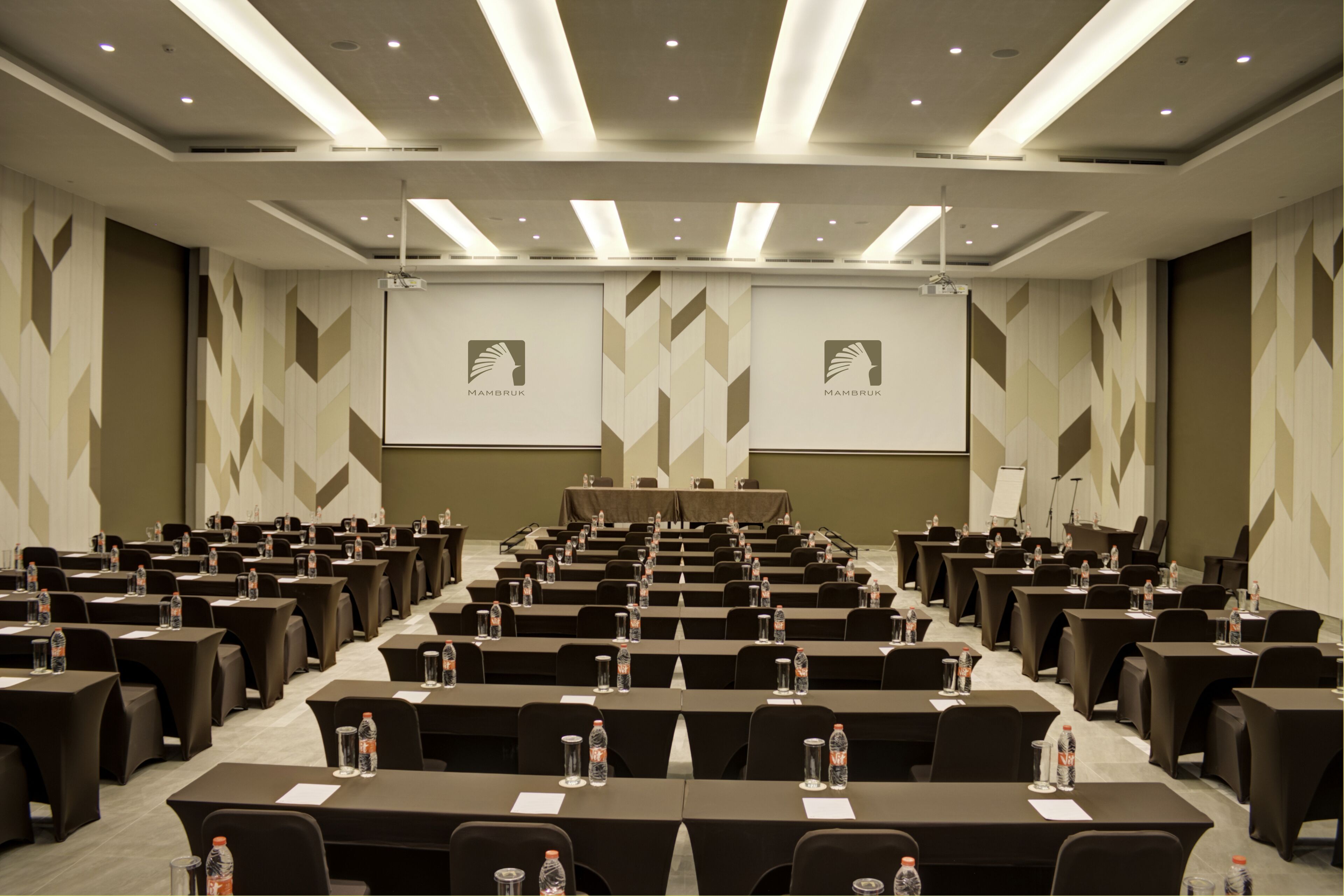 Un salón de conferencias con filas de mesas, sillas y pantallas de presentación.