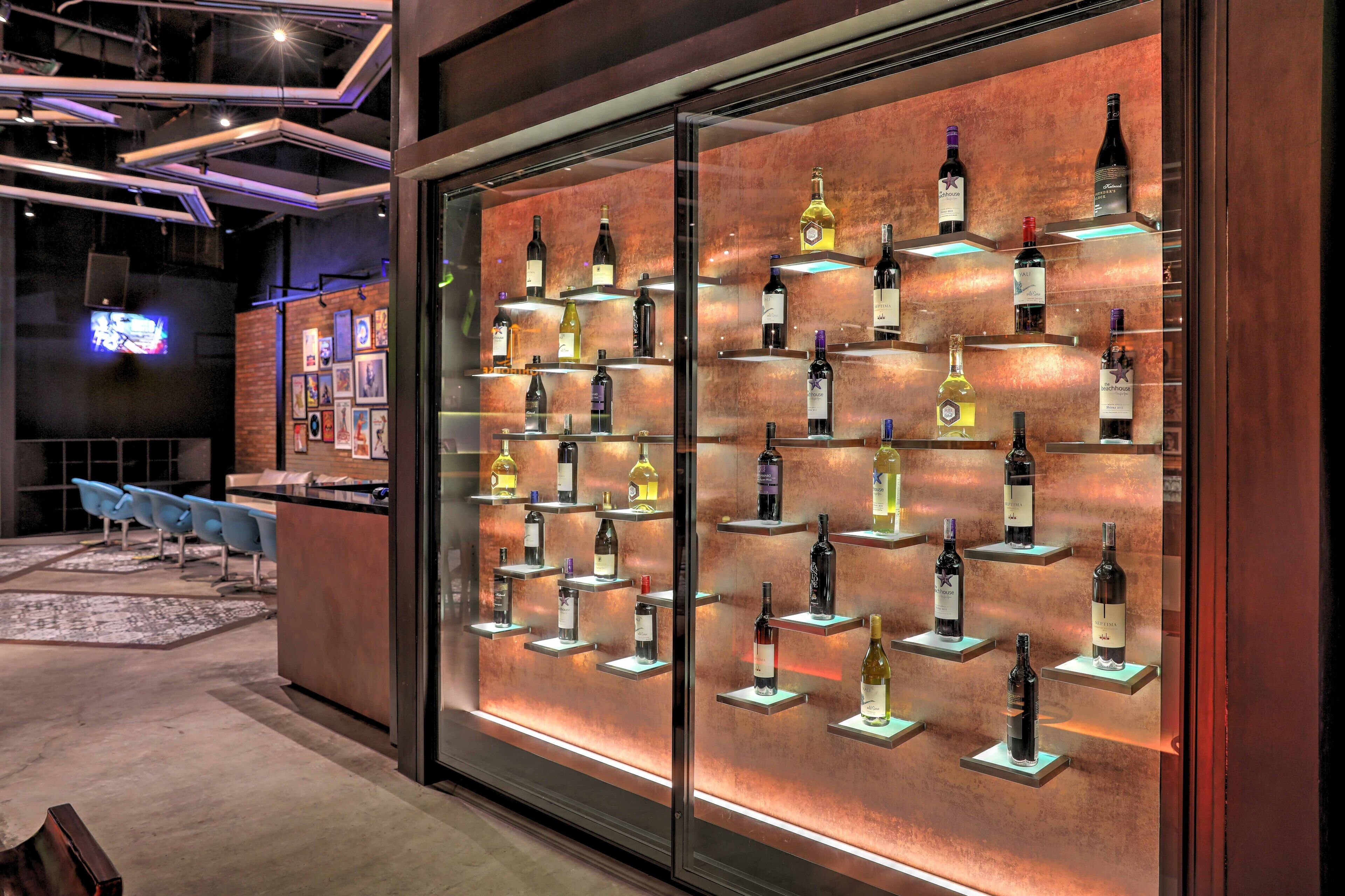 A modern bar area with multiple wine bottles displayed on shelves, a seating area with blue chairs, and a visible television screen in the background.