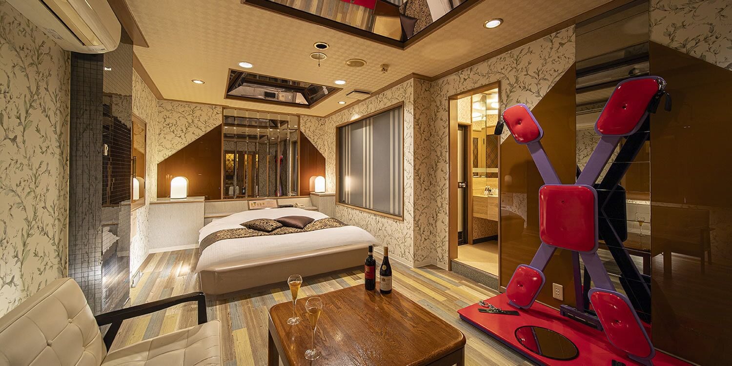 A hotel room with a bed, a wooden table with glasses, a red exercise machine, and a bathroom visible through a glass door.