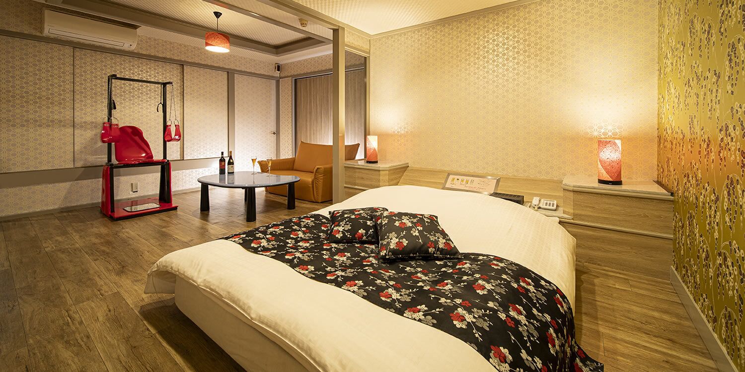A hotel room with a bed, a small table, a chair, a red coat rack, and a wall-mounted lamp.