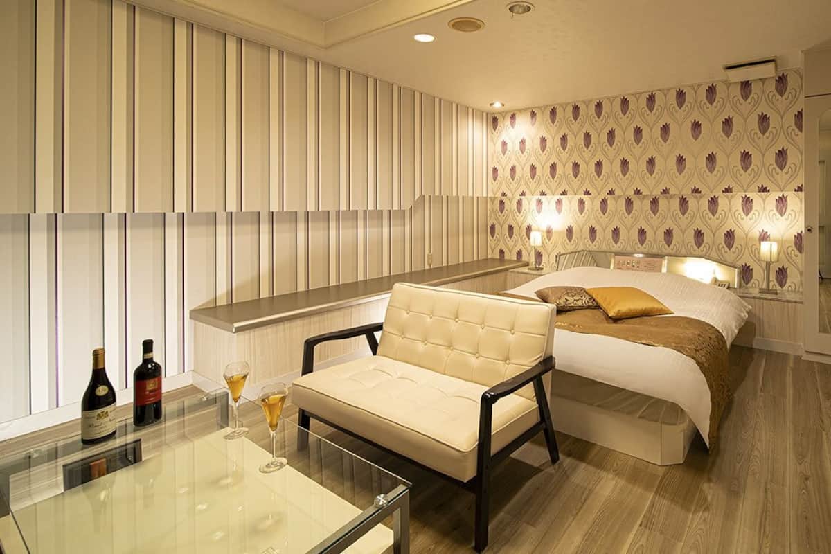 A hotel room with a bed, a sofa, a glass table with two bottles and a glass, and a patterned wallpaper.