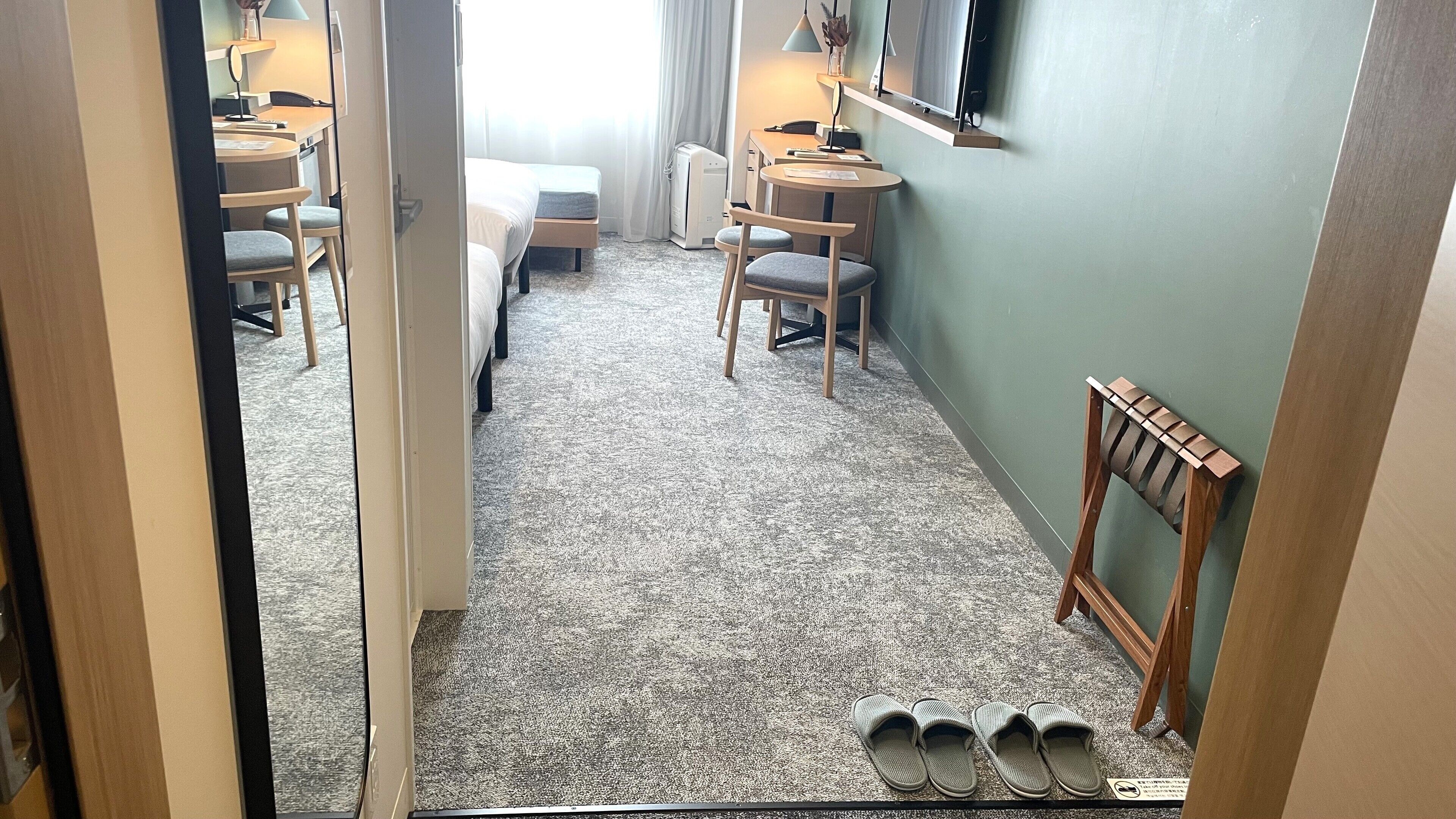 Quarto de hotel com piso carpete, uma cama, escrivaninha com cadeira, pequena mesa com cadeira, televisão na parede e sapateira.