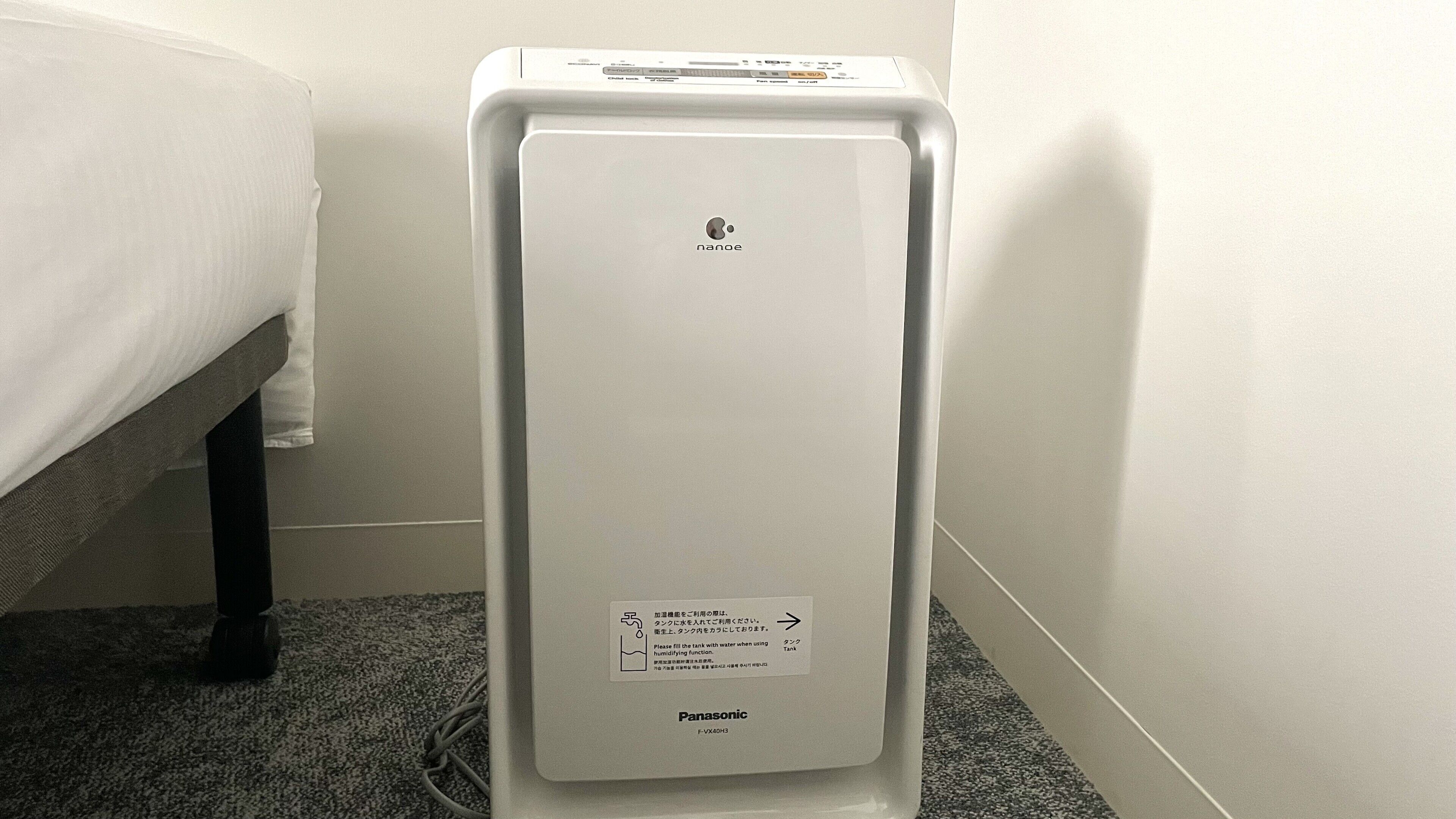 A white Panasonic humidifier with a control panel, placed in a corner of a room with a bed and a carpeted floor.