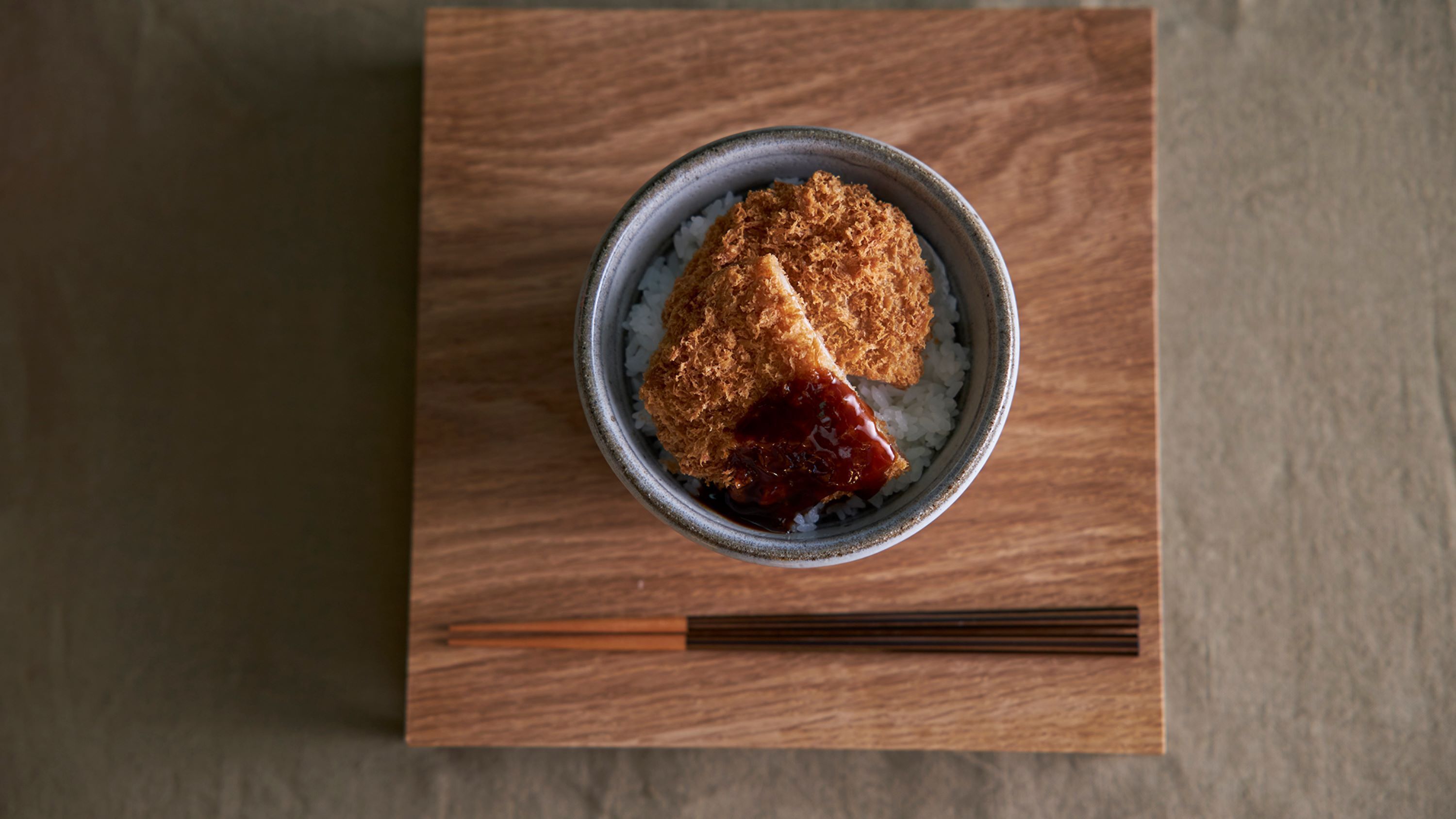 A bowl of rice topped with fried food and a dark sauce, placed on a wooden board.