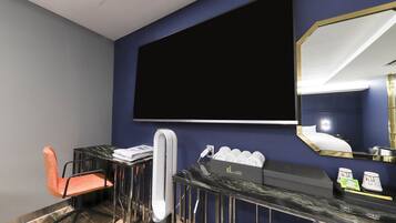 A hotel room with a large flat-screen TV mounted on the wall, a modern bathroom vanity, a chair, and a mirror.