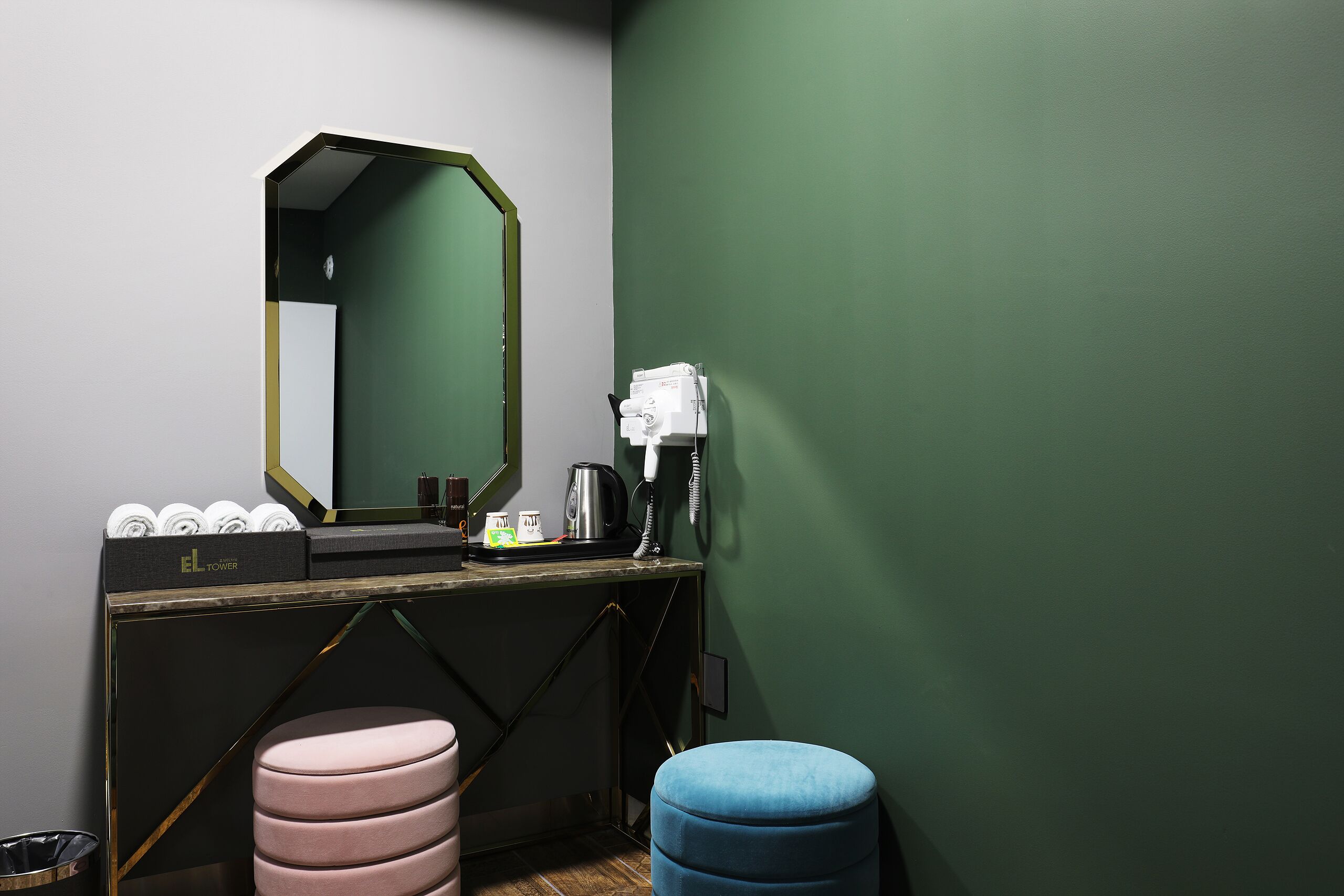 Um banheiro com parede verde, um espelho, pia com produtos de higiene, um banquinho rosa e um otomano azul.