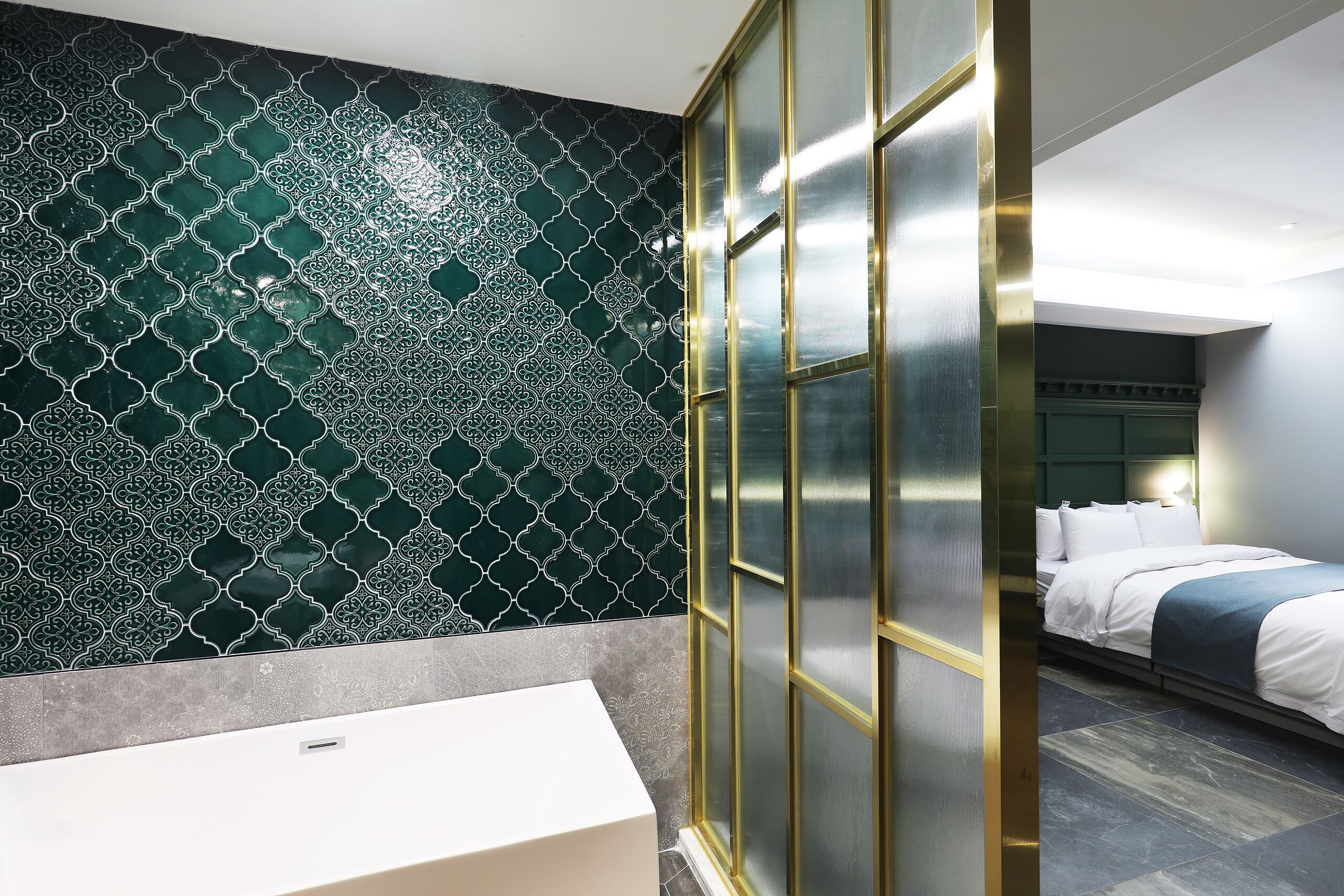 Une salle de bain moderne avec un mur recouvert de carreaux à motifs, un lavabo blanc et une porte coulissante en verre donnant sur une chambre à coucher équipée d’un lit et d’une table de chevet.