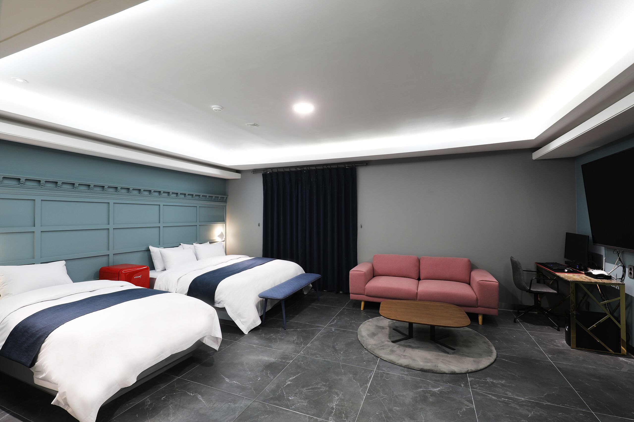 A modern hotel room with two beds, a pink sofa, a desk with a computer, and a TV.