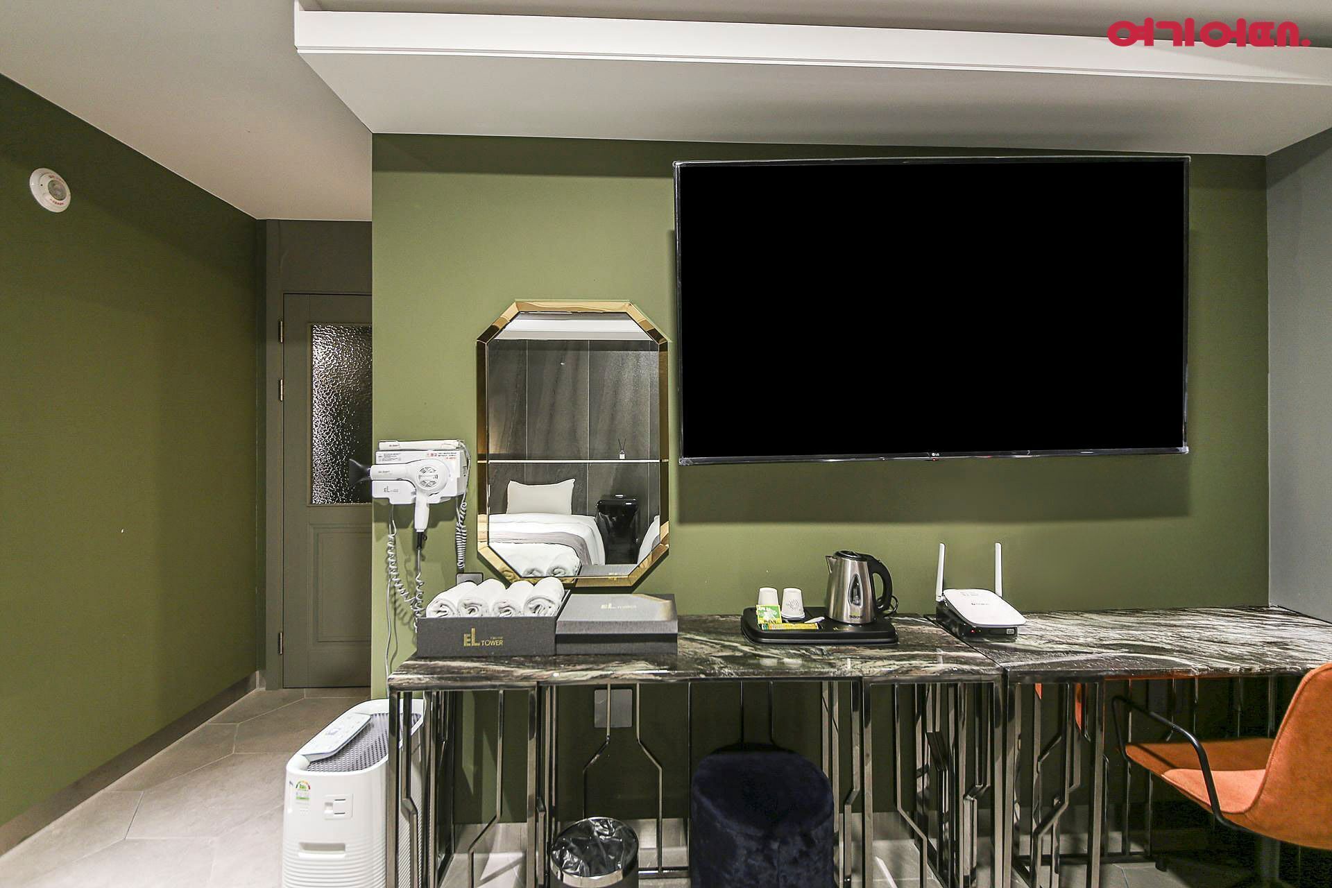 A hotel room with a large flat-screen TV mounted on the wall, a vanity with a mirror, a hairdryer, and a small table with a kettle and cups.