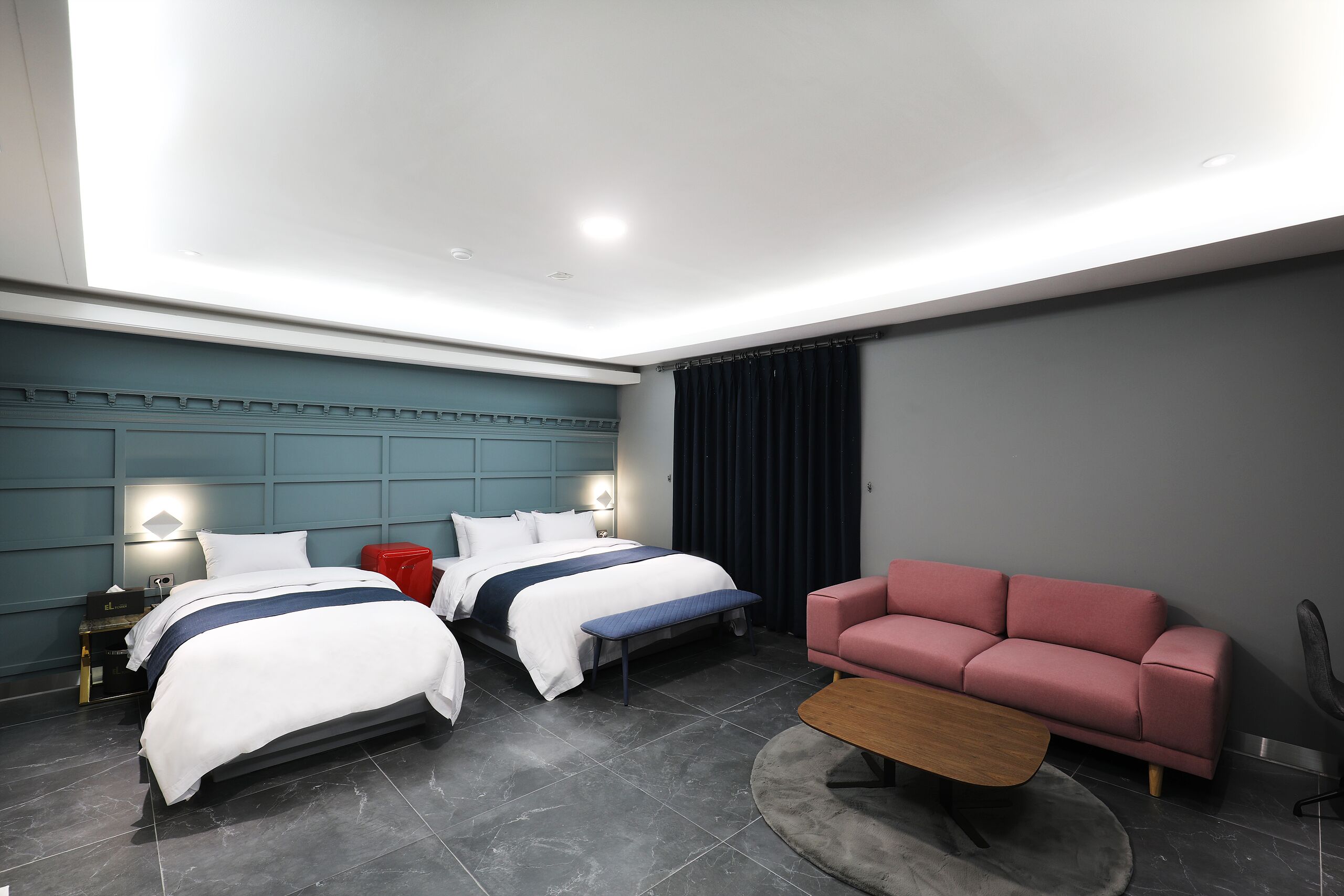 Quarto de hotel moderno com duas camas, um sofá vermelho, uma mesa de centro de madeira e um tapete cinza.