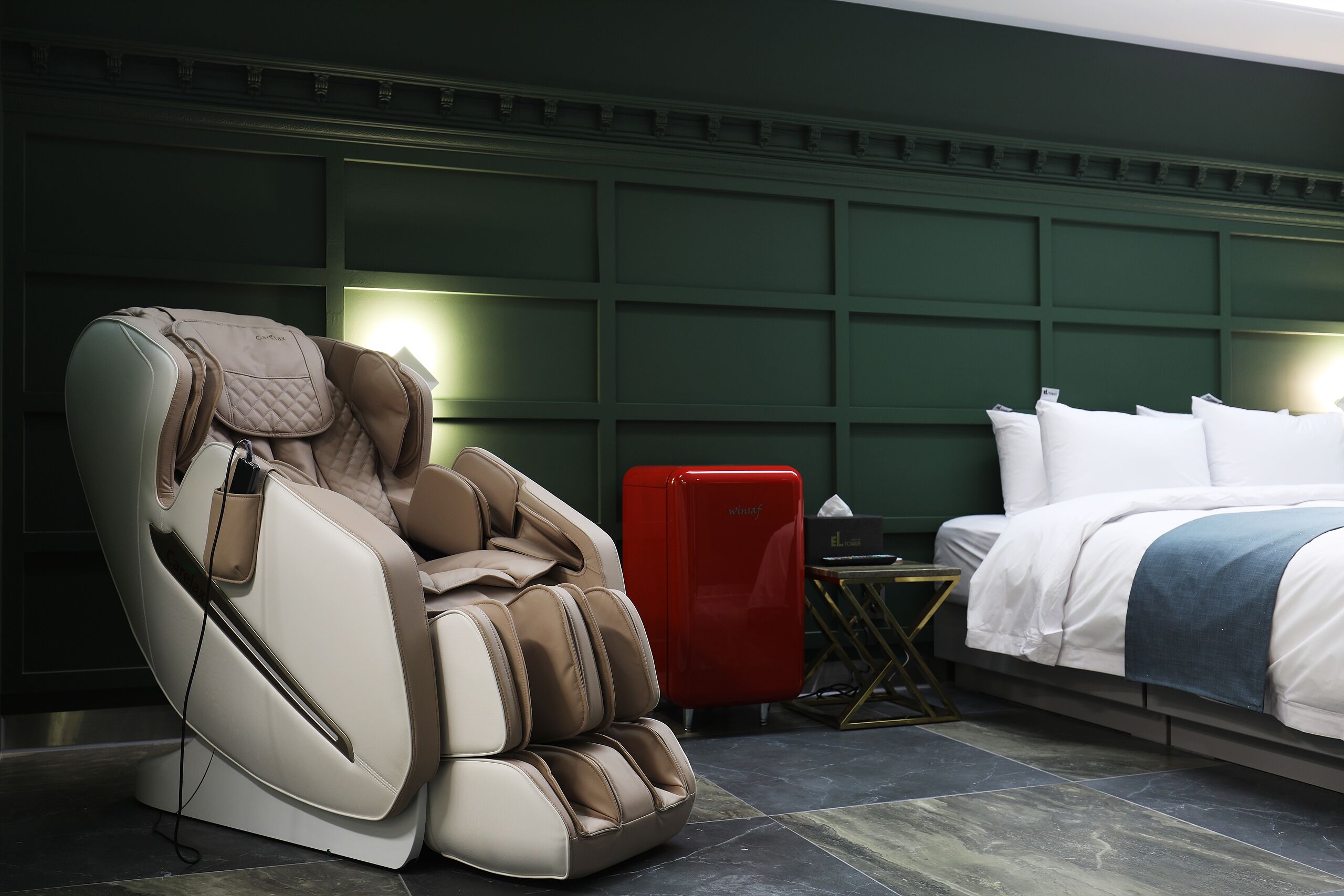 Una silla de masajes, un minibar rojo y una cama con una manta azul en una habitación de hotel.