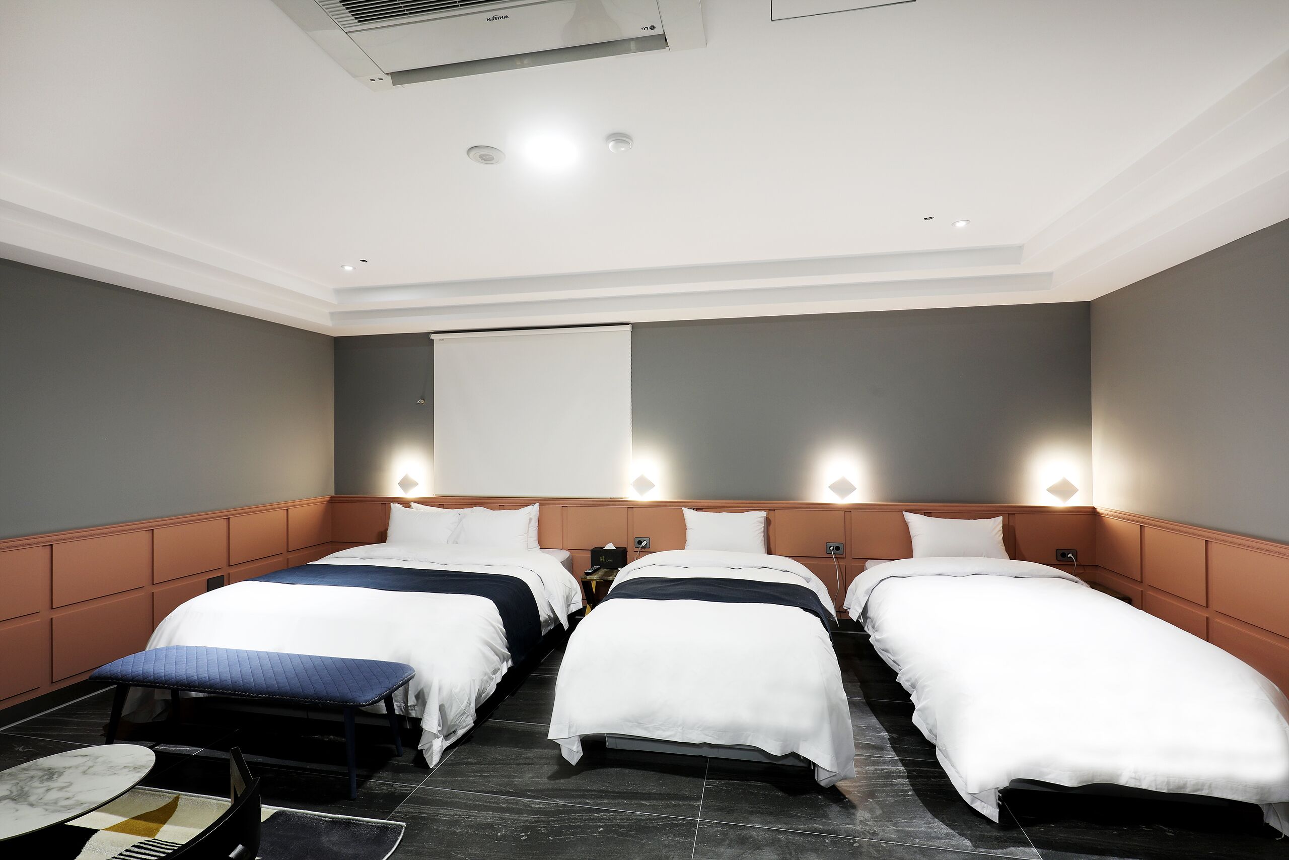 Una habitación de hotel con tres camas, una mesita pequeña y un aire acondicionado de pared.