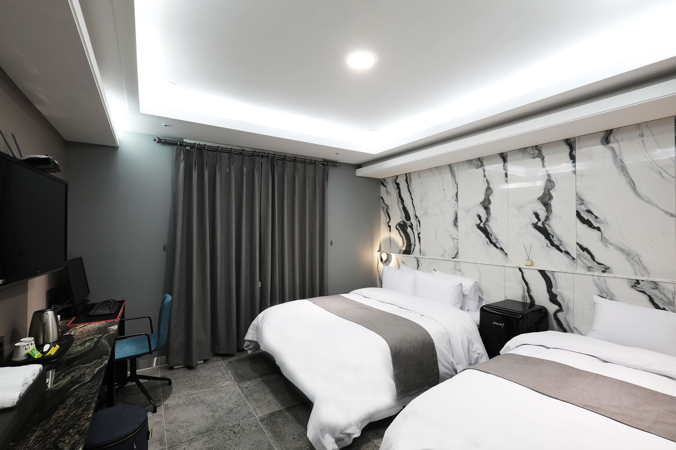 A modern hotel room with two beds, a desk with a computer, a TV, and a minibar.