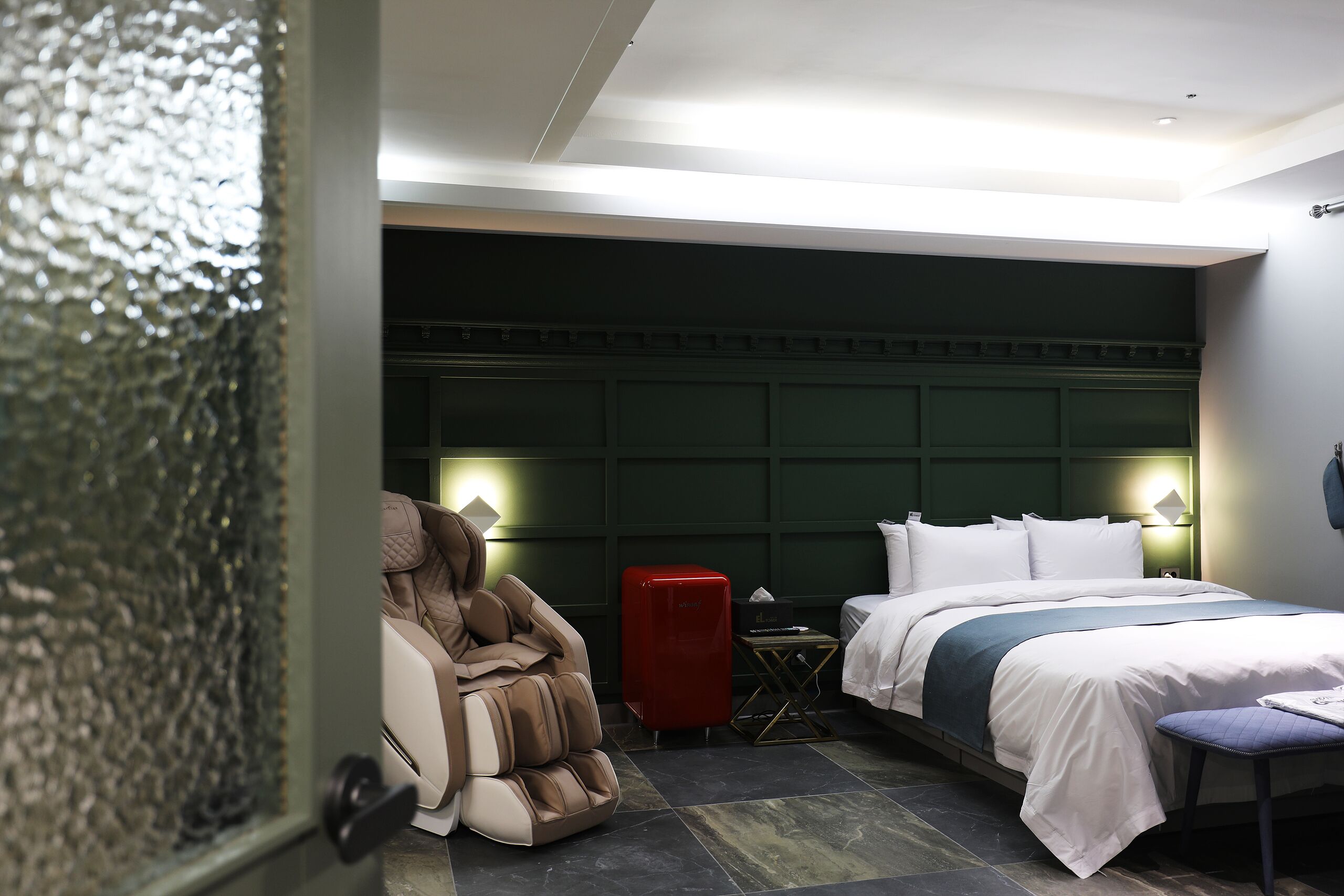 Una habitación de hotel con una cama, una silla de masaje, una maleta roja y una mesita.