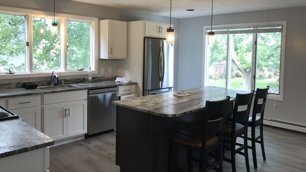 A modern kitchen with white cabinets, a stainless steel refrigerator, and a large island with bar stools. - Simple and clean living by the beach! (Narragansett)