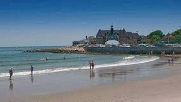 Una spiaggia con un grande edificio sullo sfondo, acqua calma e alcune persone.