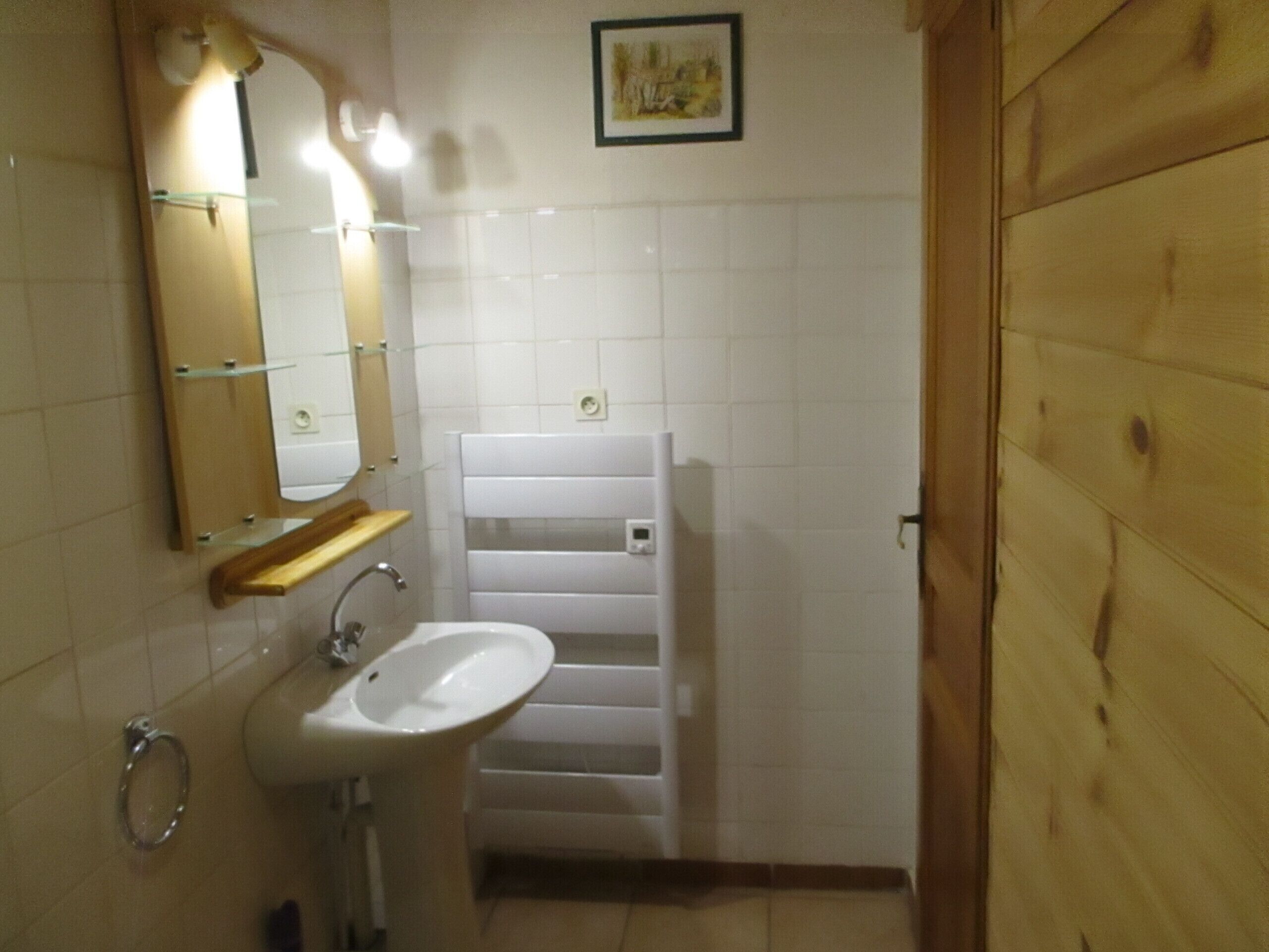 Une salle de bain avec un lavabo blanc, un miroir et des murs carrelés. Il y a une porte en bois sur la droite et un tableau encadré au mur.