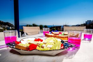 A table with a salad, sandwiches, and pink drinks, set against a scenic view.