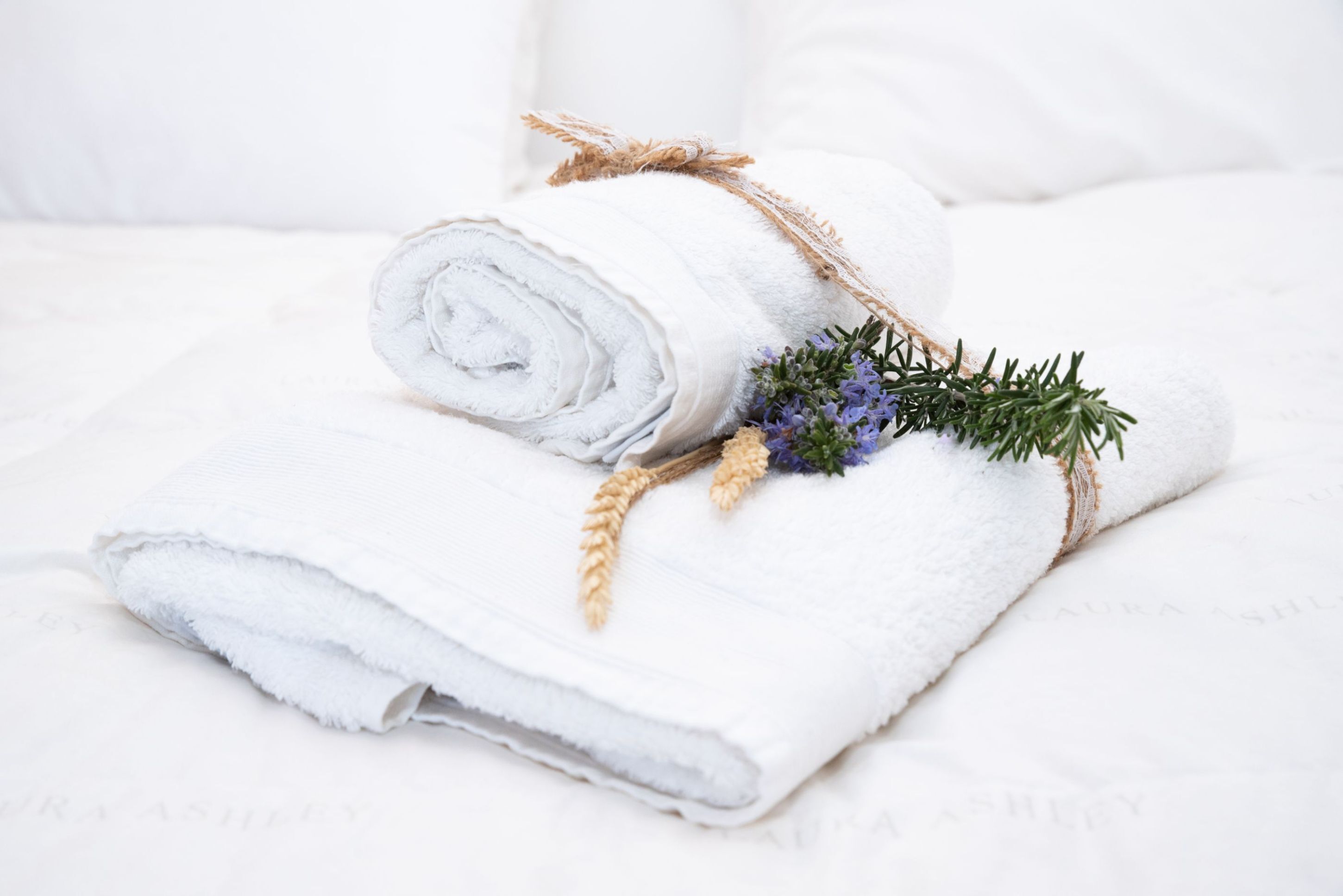 A neatly rolled white towel with a rustic twine and dried wheat, accompanied by a sprig of rosemary, placed on a white bed.