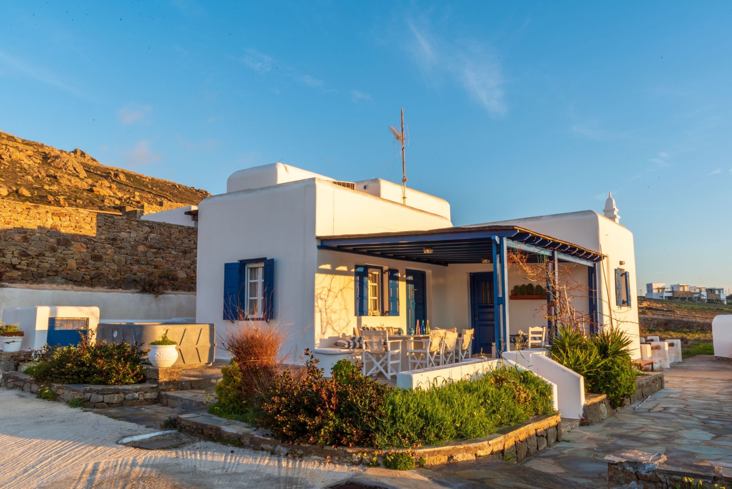A white building with blue shutters, a covered patio with outdoor furniture, and a stone wall in the background.