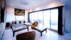 A modern living room with a sofa set, a coffee table, and a wicker chair. There is a large window with a view of the outdoors. - Indigo Geohouse DS1 (Iwami)