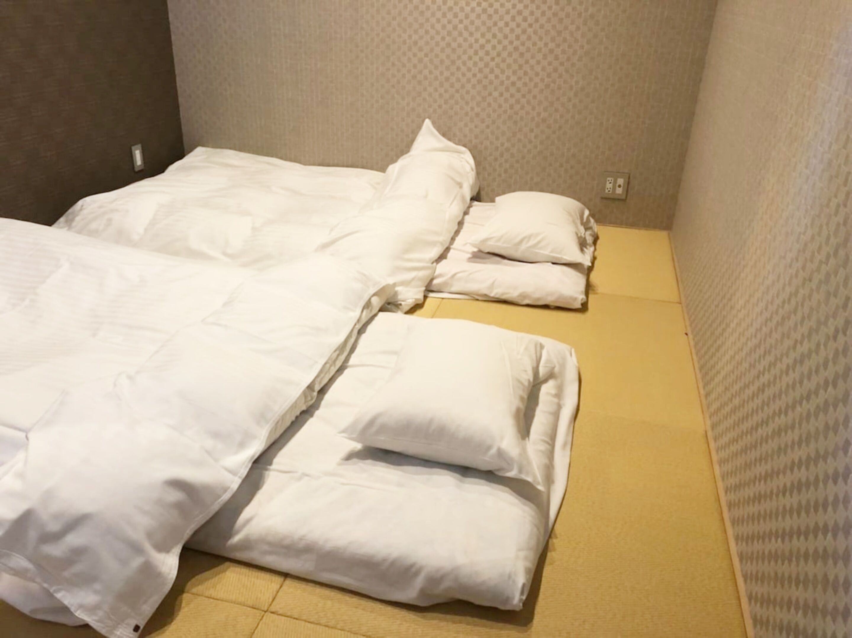 Una habitación pequeña con una cama individual, una colcha blanca sencilla, almohadas, suelo de baldosas y pared con textura.