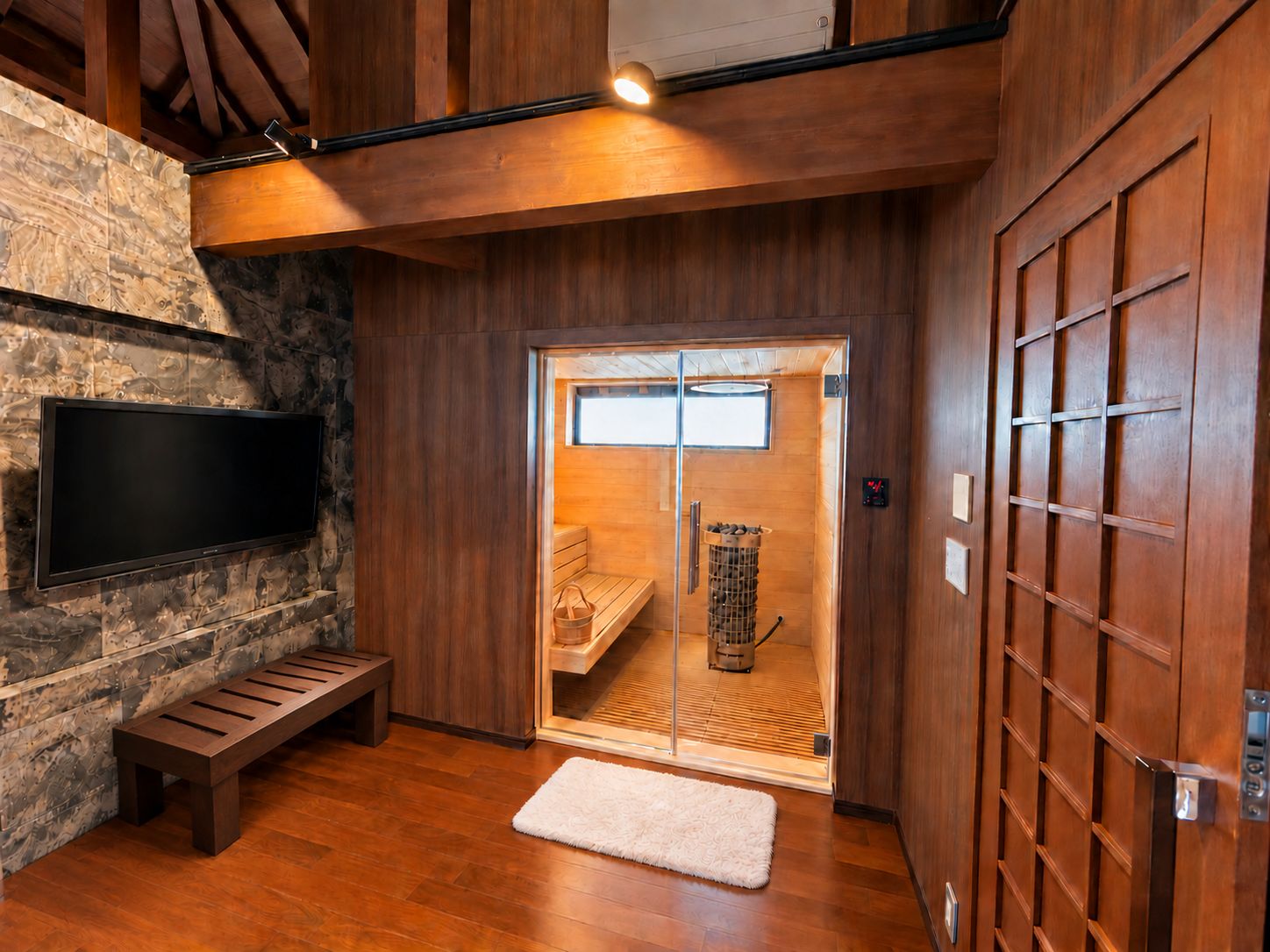 A sauna room with a wooden bench, a television mounted on a stone wall, and a sliding glass door leading to a sauna area.