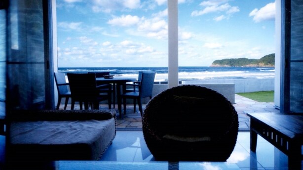 A room with a large window overlooking the ocean, a seating area with a table and chairs, and a round, textured ottoman. - Indigo Geohouse DS1 (Iwami)