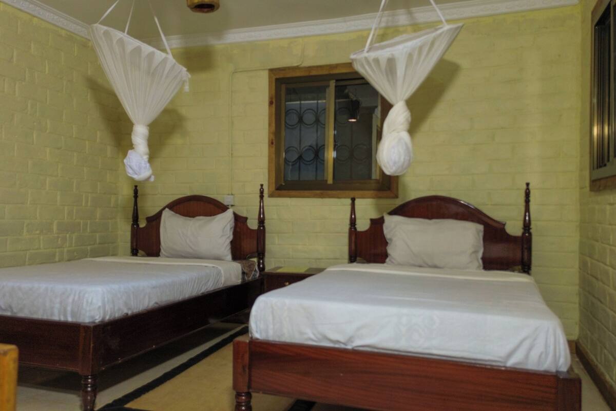 Two wooden beds with white linens, a window with a metal grille, and a hanging mosquito net above each bed.