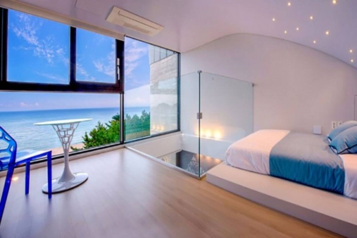 A modern bedroom with a large window offering a view of the sea, a glass partition, and a bed with blue bedding.