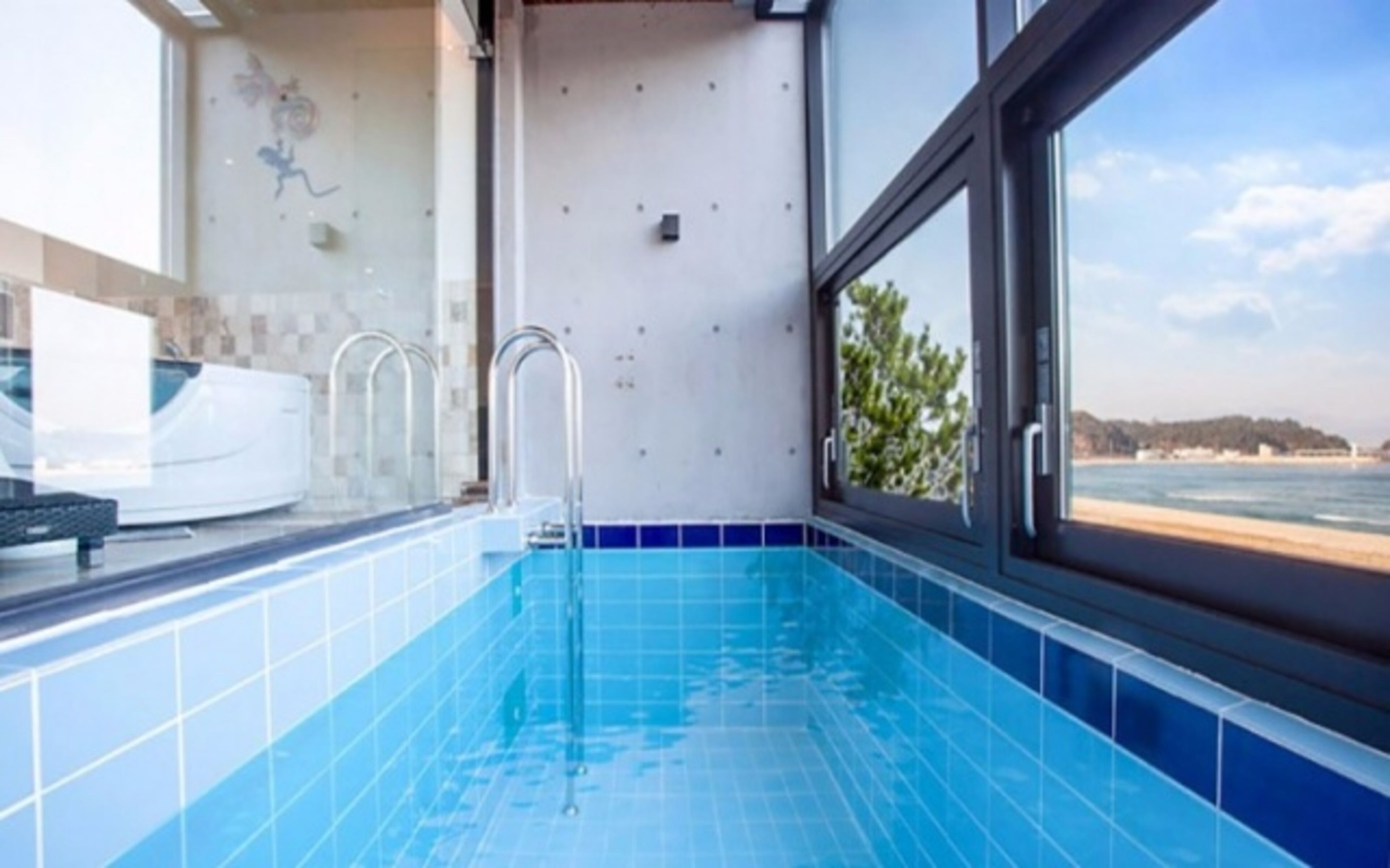 Ein moderner Innenpool mit hellblauen Fliesen, einem Fenster mit Meerblick und einer weißen Badewanne.