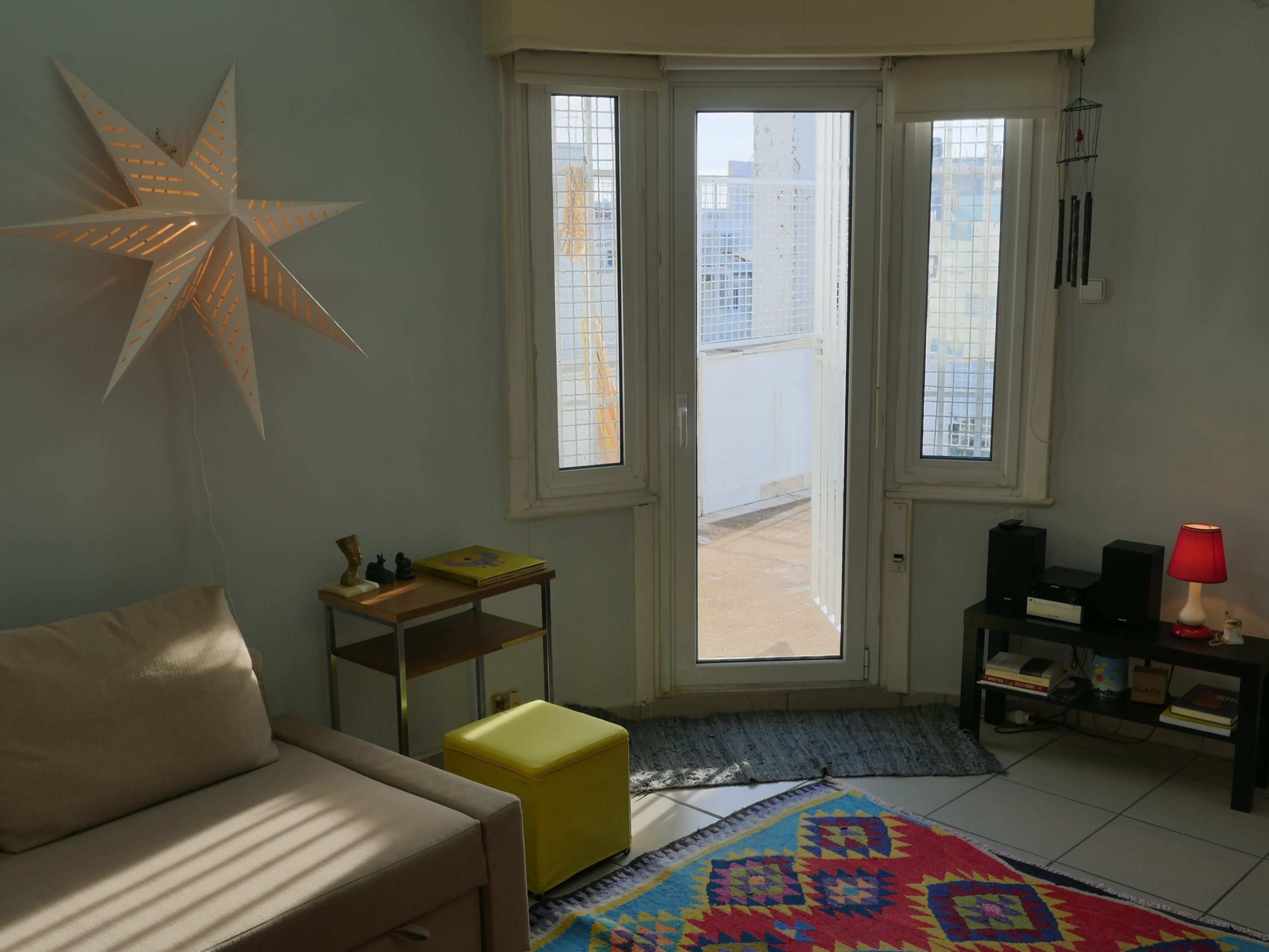 Une pièce avec un canapé, une petite table sur laquelle se trouve un livre, un tapis coloré, un luminaire en forme d’étoile et une porte donnant sur l’extérieur.