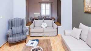 A living room with a striped armchair, a beige sofa, and a white sofa. There is a dining table with chairs and a chandelier. - The Muses House (Athens)