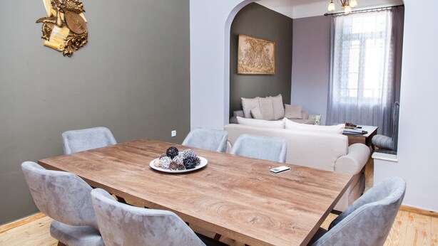 A dining area with a wooden table and grey upholstered chairs, a decorative wall piece, and a cozy seating area with a sofa and a painting. - The Muses House (Athens)