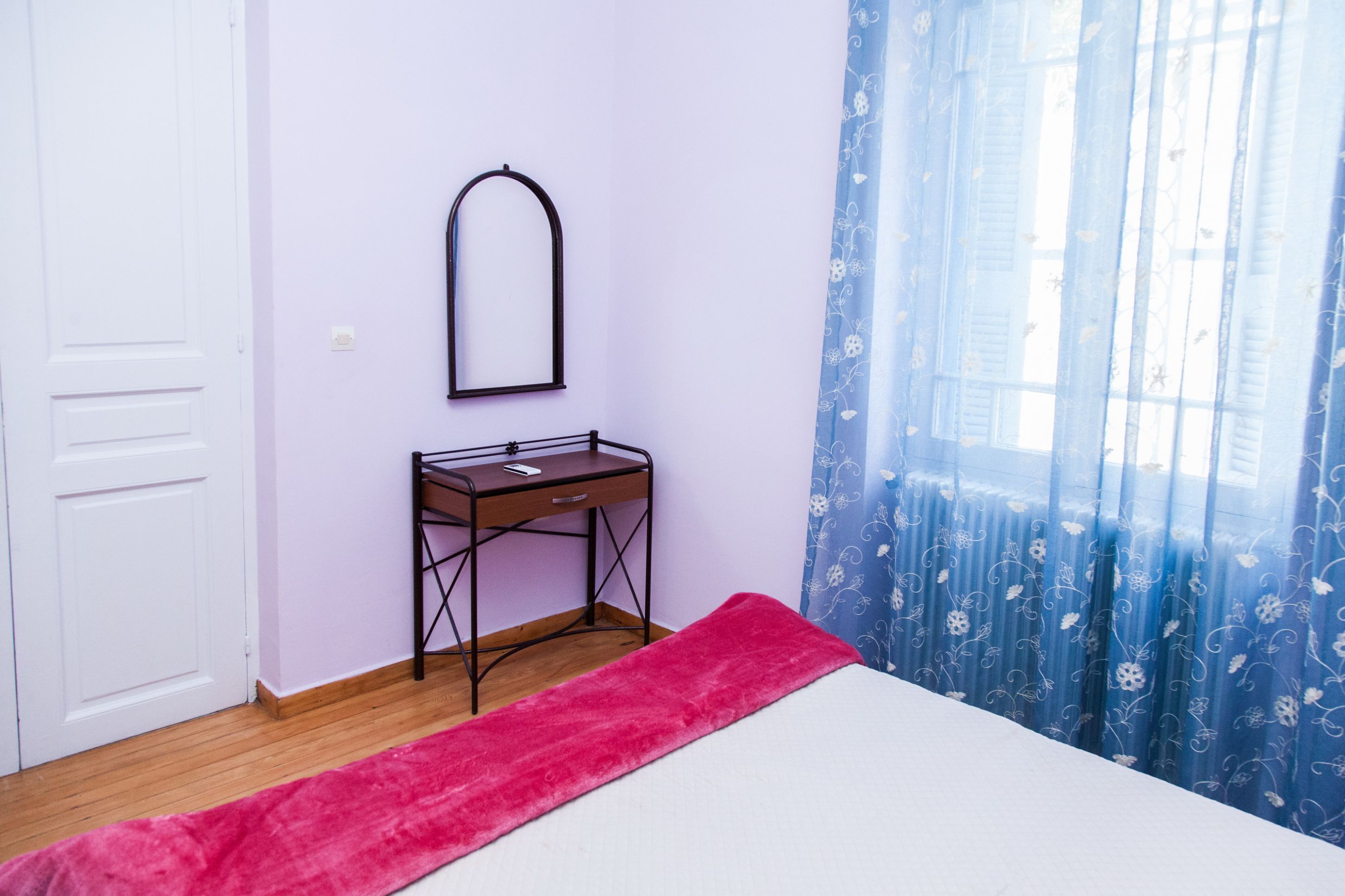 A bedroom with a bed, a desk, a mirror, and a window with curtains.