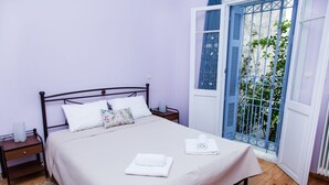 A neatly made bed with a white bedspread and two folded towels, a nightstand with a lamp, and a view of a balcony with blue shutters. - The Muses House (Athens)