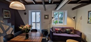 A cozy living room with a wooden table, purple sofa, and two windows providing natural light.