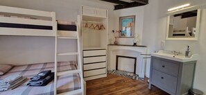 A bunk bed with a checkered bedspread, a white wardrobe, a fireplace with a vase of flowers, and a bathroom with a sink and mirror.