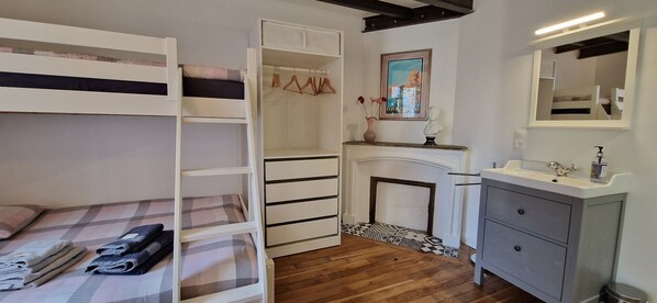 A bunk bed with a checkered bedspread, a white wardrobe, a fireplace with a vase of flowers, and a bathroom with a sink and mirror.